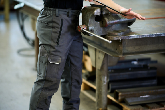 Arbeiter in einer Getzner Textil Arbeitshose aus strapazierfähigem Material, bedient einen Schraubstock in einer Werkstatt, was die Funktionalität und Robustheit der Berufsbekleidung unterstreicht.