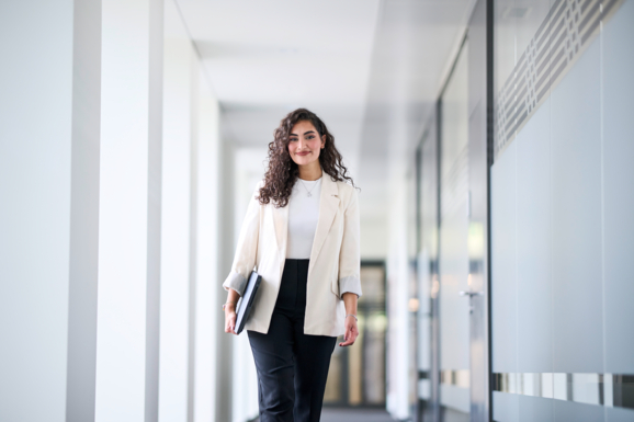 Eda Kurt hat ein Notebook in der Hand und läuft lächelnd einen lichtdurchfluteten Gang entlang.
