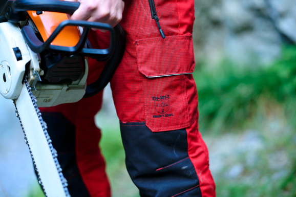 Nahaufnahme einer robusten, roten Arbeitshose von Getzner Textil, die Sicherheitsstandards erfüllt, getragen von einem Profi mit einer Motorsäge in der Hand, um die Langlebigkeit und Qualität der Schutzbekleidung zu demonstrieren.