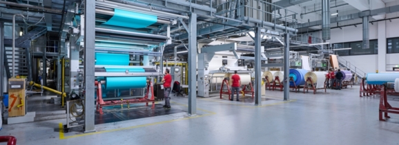 Eine große Maschinenhalle, in der ein Spannrahmen zu sehen ist, in dem gerade ein türkiser Stoff eingespannt ist.