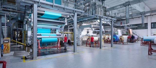 Eine große Maschinenhalle, in der ein Spannrahmen zu sehen ist, in dem gerade ein türkiser Stoff eingespannt ist.