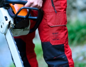 Nahaufnahme einer robusten, roten Arbeitshose von Getzner Textil, die Sicherheitsstandards erfüllt, getragen von einem Profi mit einer Motorsäge in der Hand, um die Langlebigkeit und Qualität der Schutzbekleidung zu demonstrieren.
