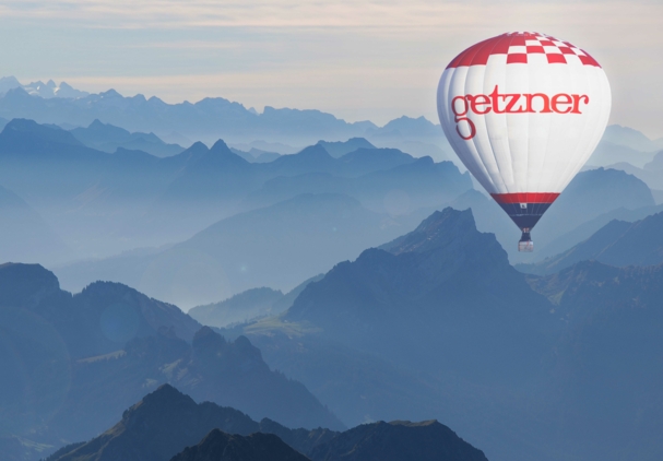Heißluftballon mit Getzner Textil Logo schwebt über einer Bergkette unter klarem blauen Himmel.