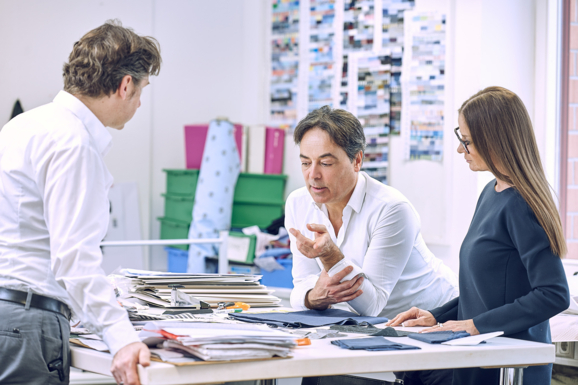 Drei Mode-Designer diskutieren über ein Projekt in ihrem hellen Atelier, umgeben von Stoffproben, Skizzen und Arbeitsmaterialien.
