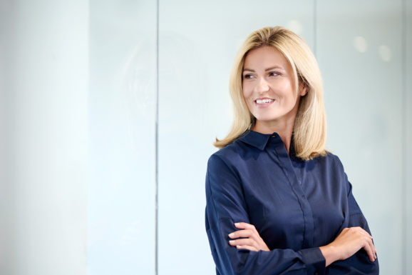 Eine selbstbewusste Geschäftsfrau in einer dunkelblauen Corporate Fashion Bluse von Getzner Textil lächelt, während sie in einem modernen Büro steht.
