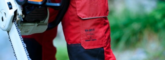 Nahaufnahme einer robusten, roten Arbeitshose von Getzner Textil, die Sicherheitsstandards erfüllt, getragen von einem Profi mit einer Motorsäge in der Hand, um die Langlebigkeit und Qualität der Schutzbekleidung zu demonstrieren.