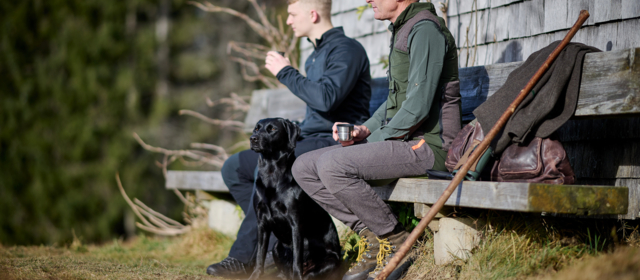 Zwei Jäger in Jagdbekleidung von Getzner Textil entspannen auf einer Holzbank und genießen die Ruhe der Natur bei einer Tasse Tee.