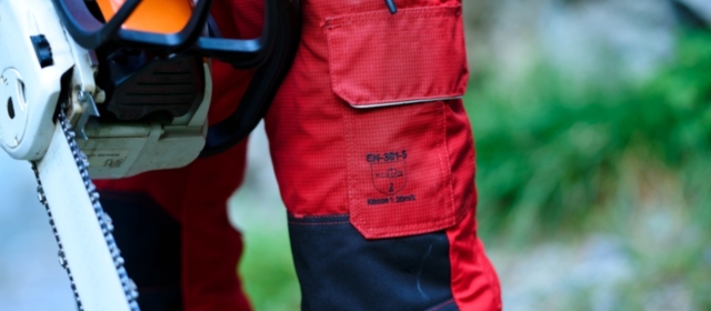 Nahaufnahme einer robusten, roten Arbeitshose von Getzner Textil, die Sicherheitsstandards erfüllt, getragen von einem Profi mit einer Motorsäge in der Hand, um die Langlebigkeit und Qualität der Schutzbekleidung zu demonstrieren.